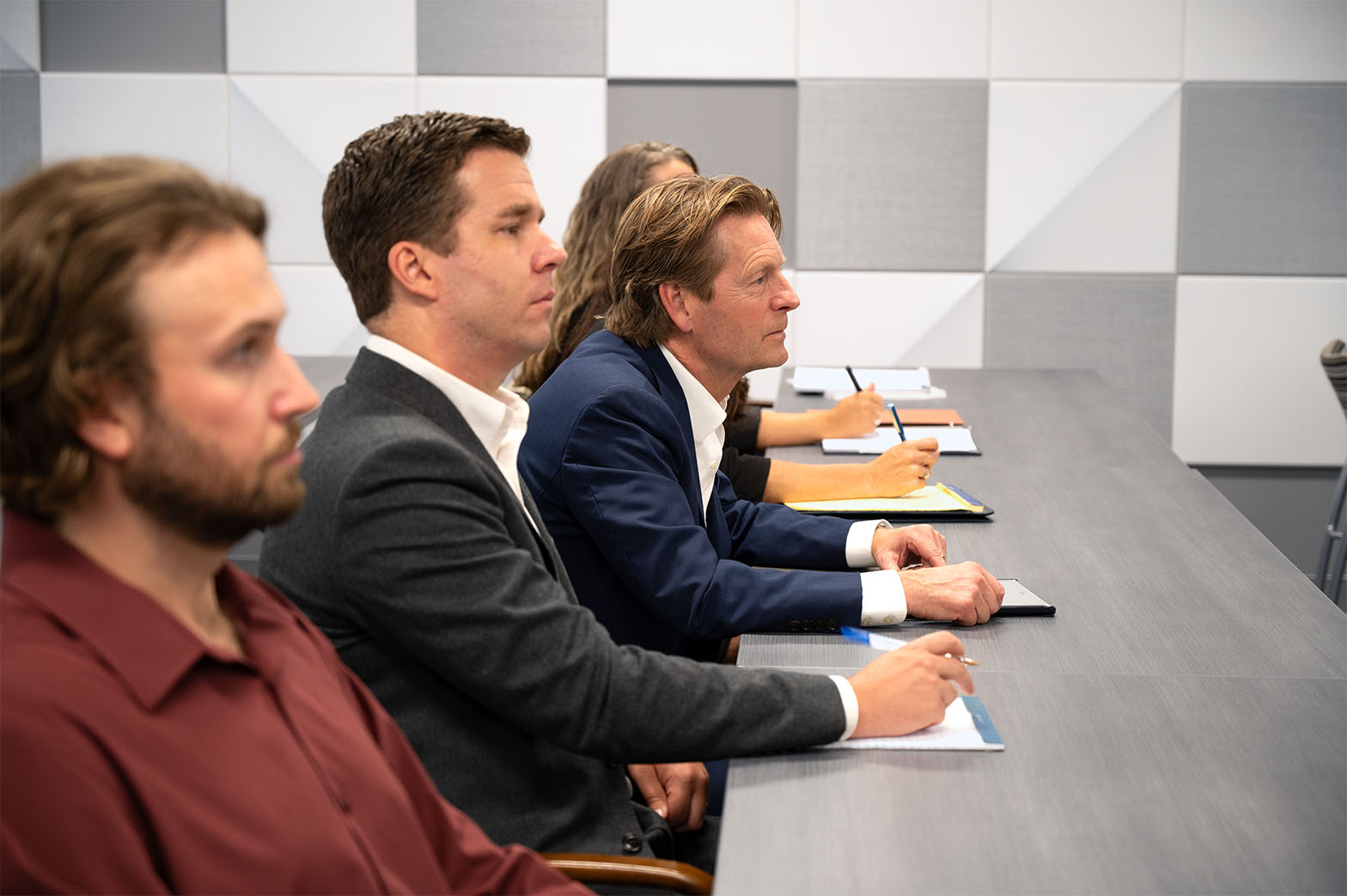 Attendees seated in a row at a conference table, listening and taking notes in a modern office.