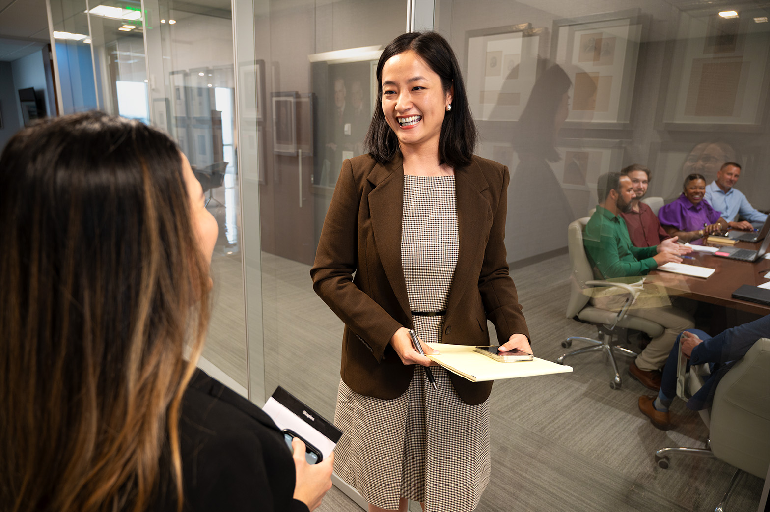 Smiling professional woman speaking with a colleague as a meeting happens behind her.